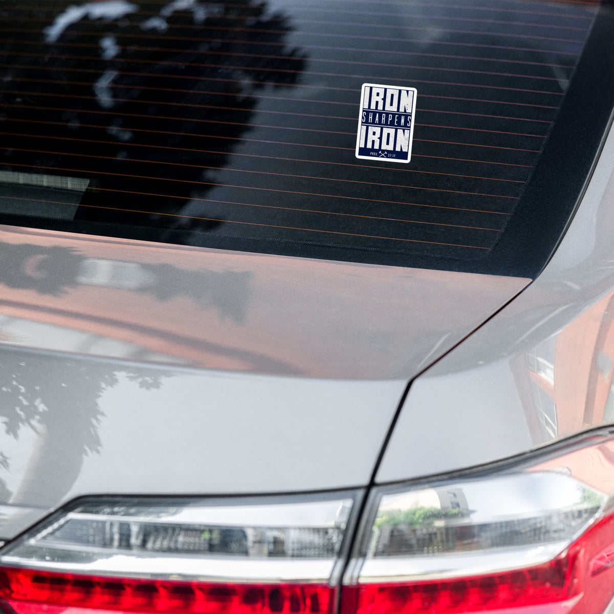 A rectangular vinyl sticker with the text 'IRON SHARPENS IRON' in white on a navy blue background, applied to the rear window of a silver car.