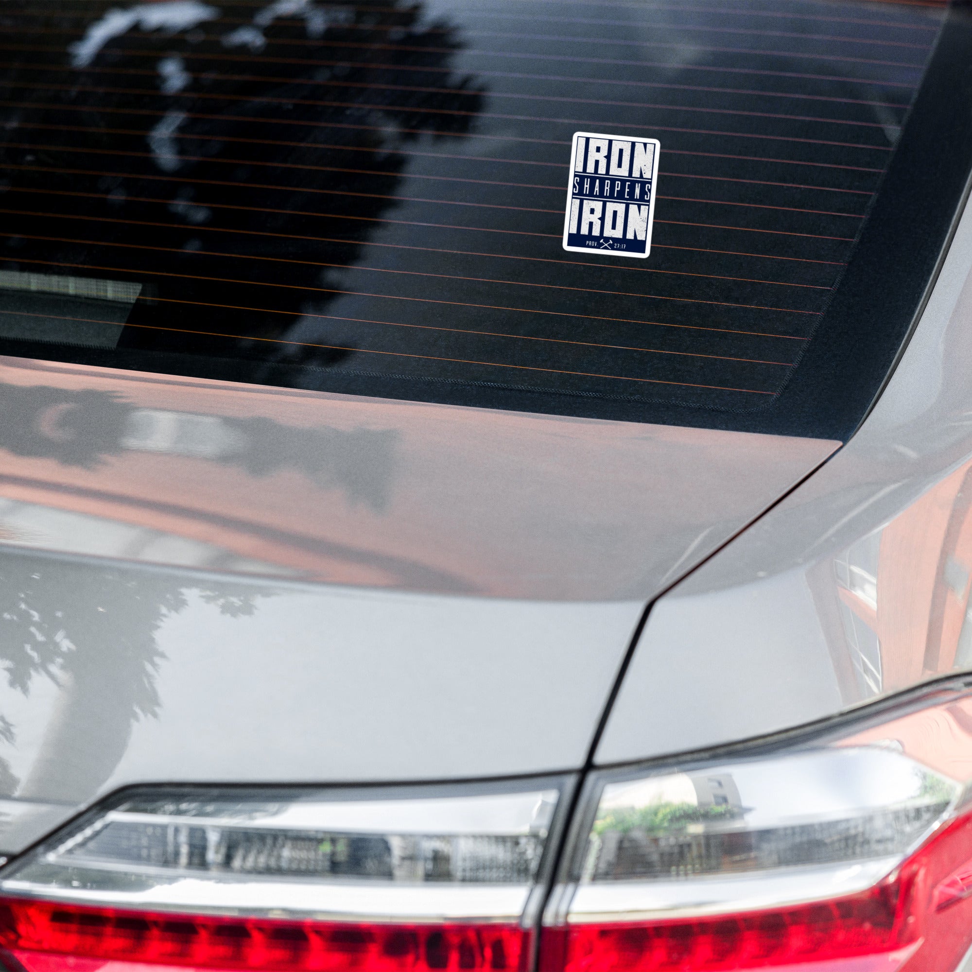 A rectangular vinyl sticker with the text 'IRON SHARPENS IRON' in white on a navy blue background, applied to the rear window of a silver car.
