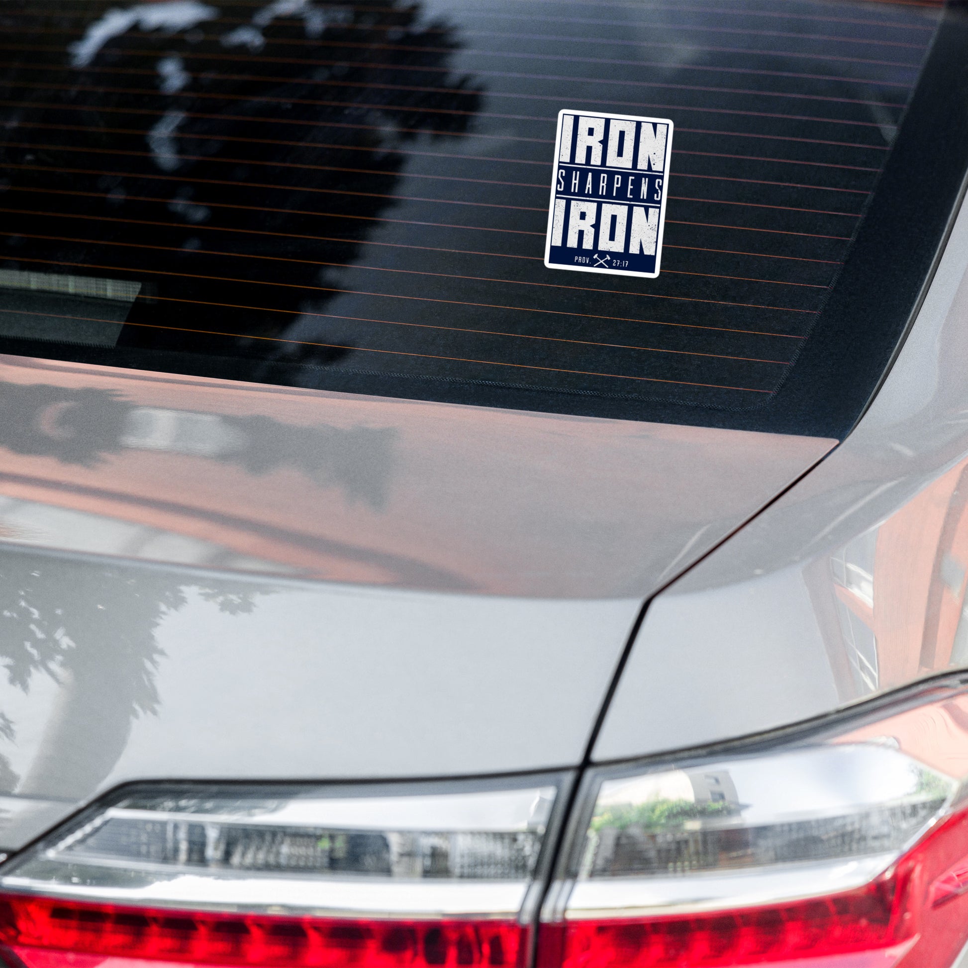 A rectangular vinyl sticker with the text 'IRON SHARPENS IRON' in white on a navy blue background, applied to the rear window of a silver car.