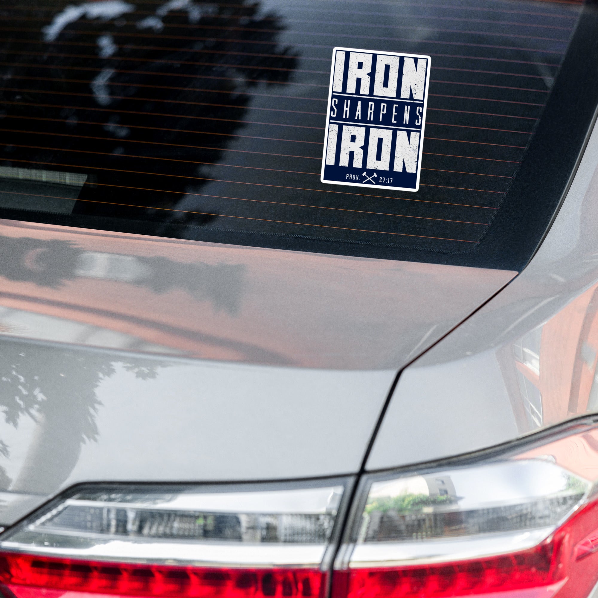 A rectangular vinyl sticker with the text 'IRON SHARPENS IRON' in white on a navy blue background, applied to the rear window of a silver car.